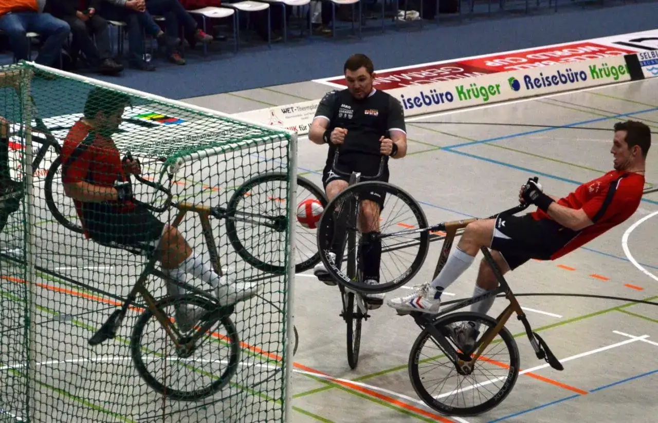 Zawodnicy grają w piłkę rowerową, próbując zdobyć bramkę. Jeden z nich próbuje uderzyć piłkę nogą, siedząc na rowerze.