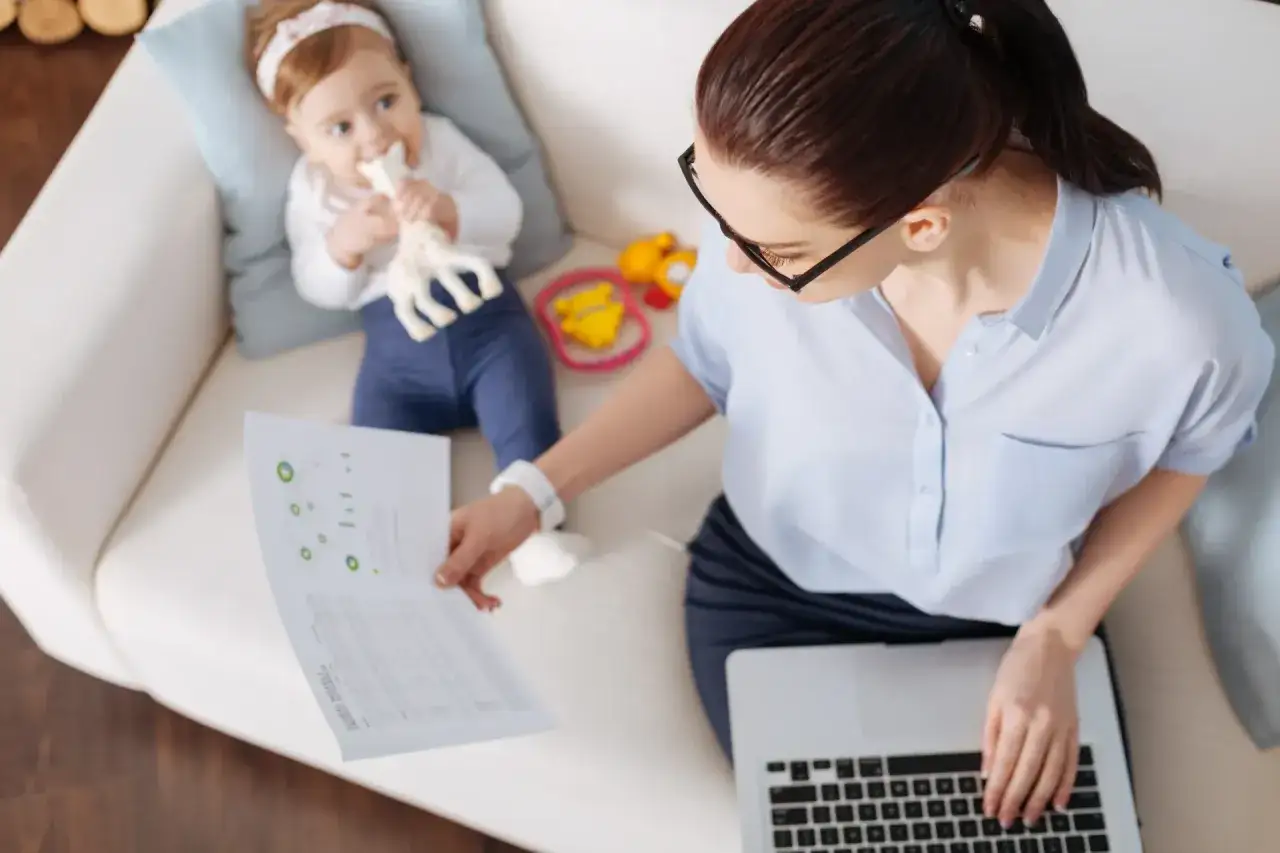 Kobieta pracuje na laptopie, podczas gdy dziecko bawi się na kanapie. To obrazek idealnie ilustrujący wyzwania związane z dyrektywą work life balance.