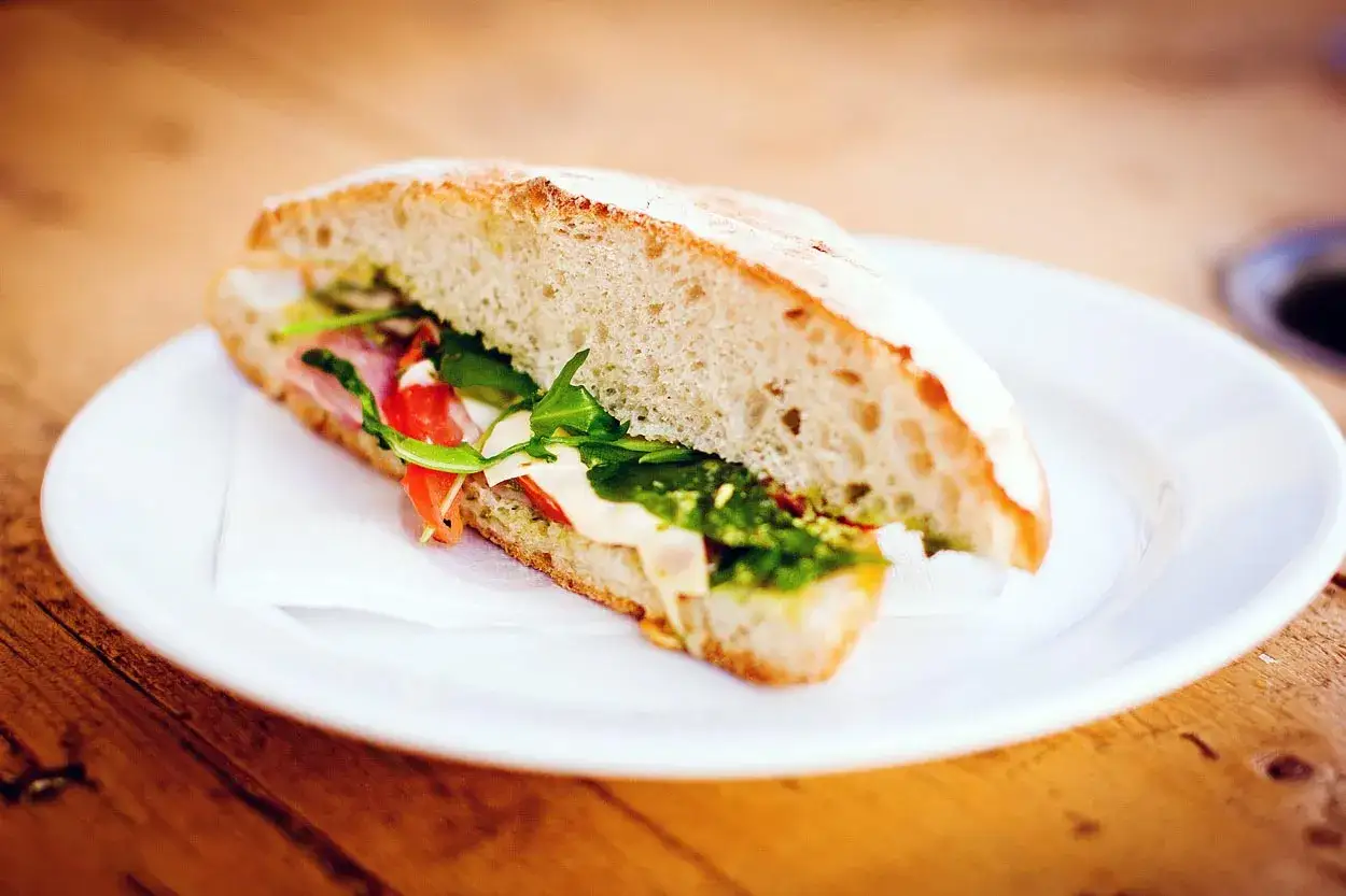Un delicioso sandwich de hummus con jamón, rúcula y tomate, servido en pan rústico sobre un plato blanco.