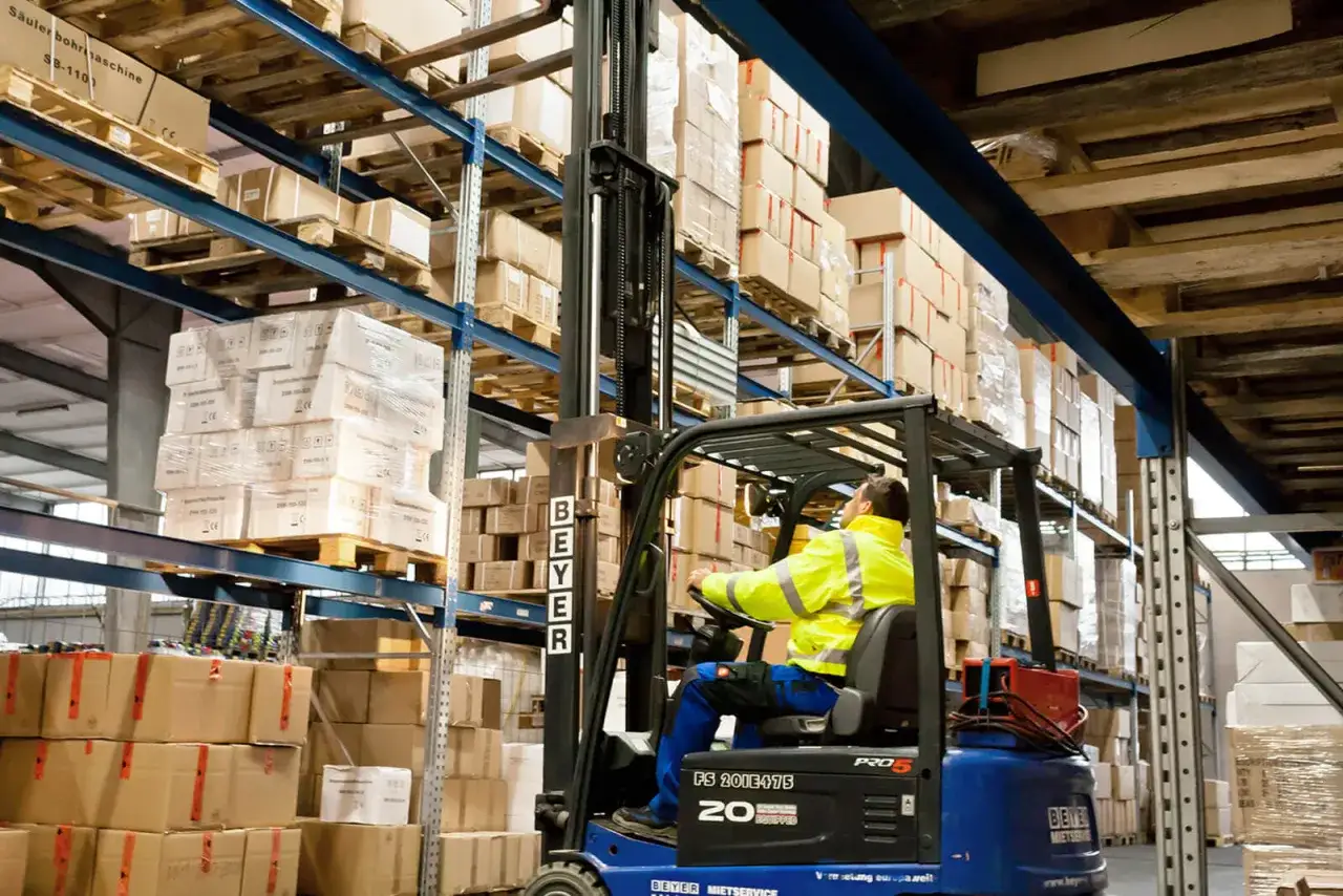 Gabelstaplerfahrer in gelber Warnweste bei der Logistik Arbeit im Lager, viele Kartons auf Regalen.