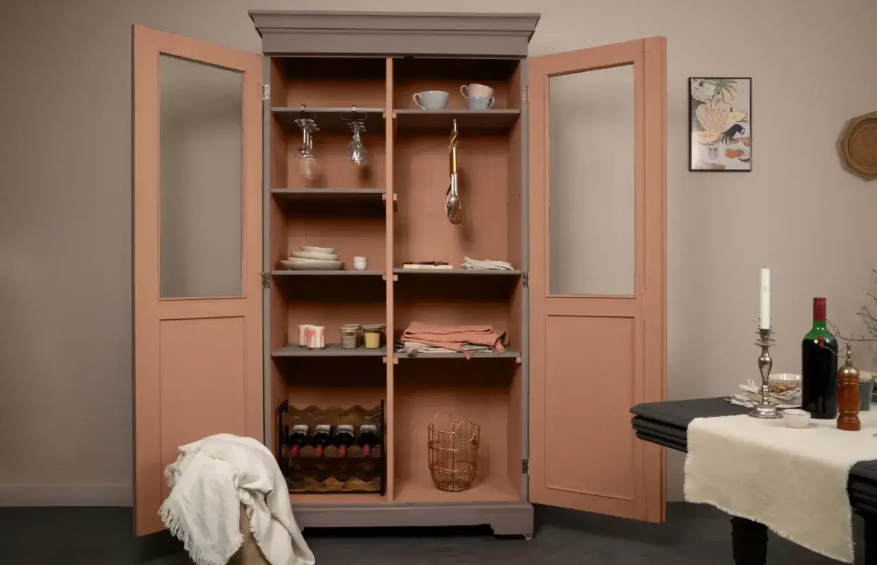Armoire vitrée avec un relooking meuble 2 couleurs : gris anthracite et rose poudré. Elle est garnie de vaisselle, verres, et bouteilles de vin.