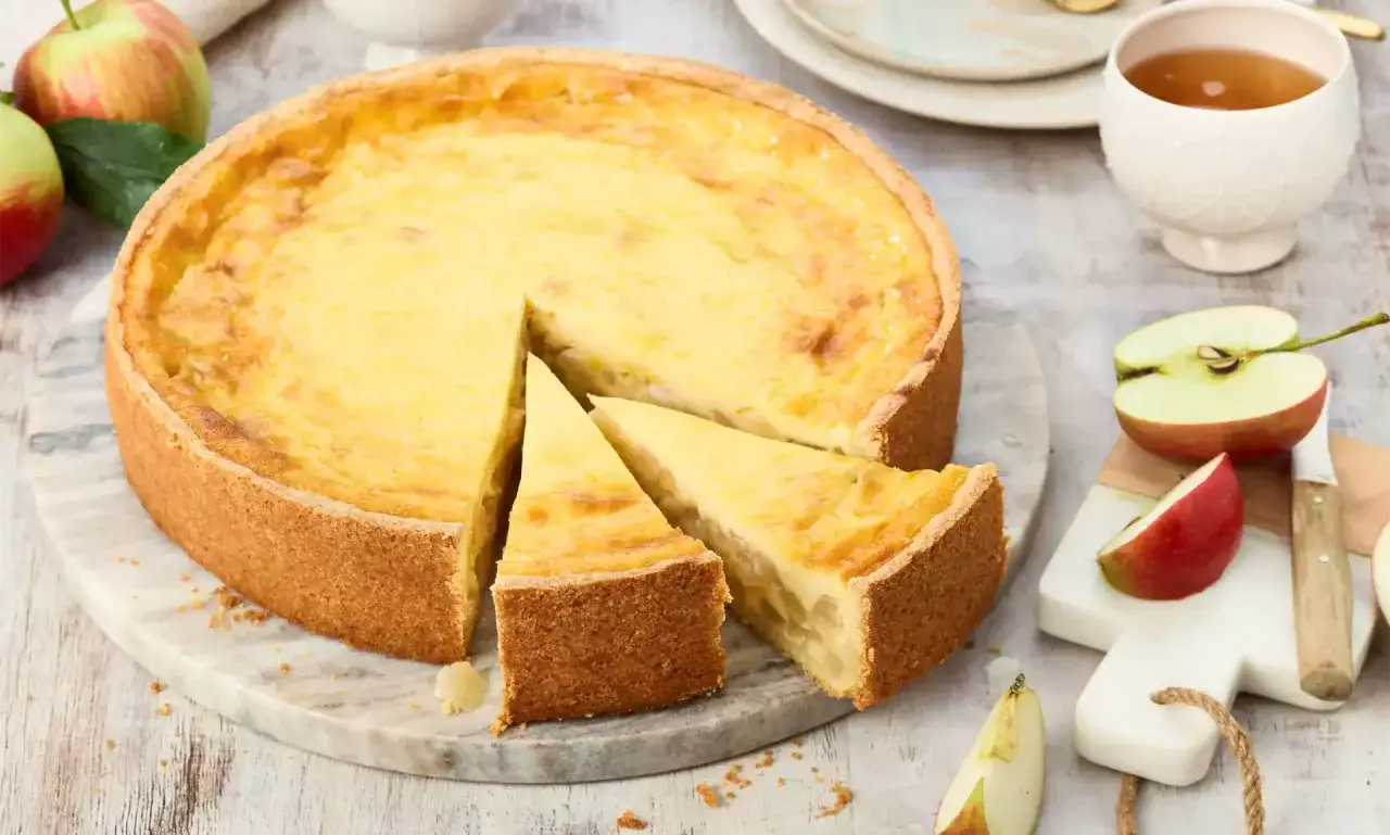 Ein saftiger Apfel Schmand Kuchen mit goldbraunem Boden und cremigem Belag, daneben frische Äpfel und eine Tasse Tee.