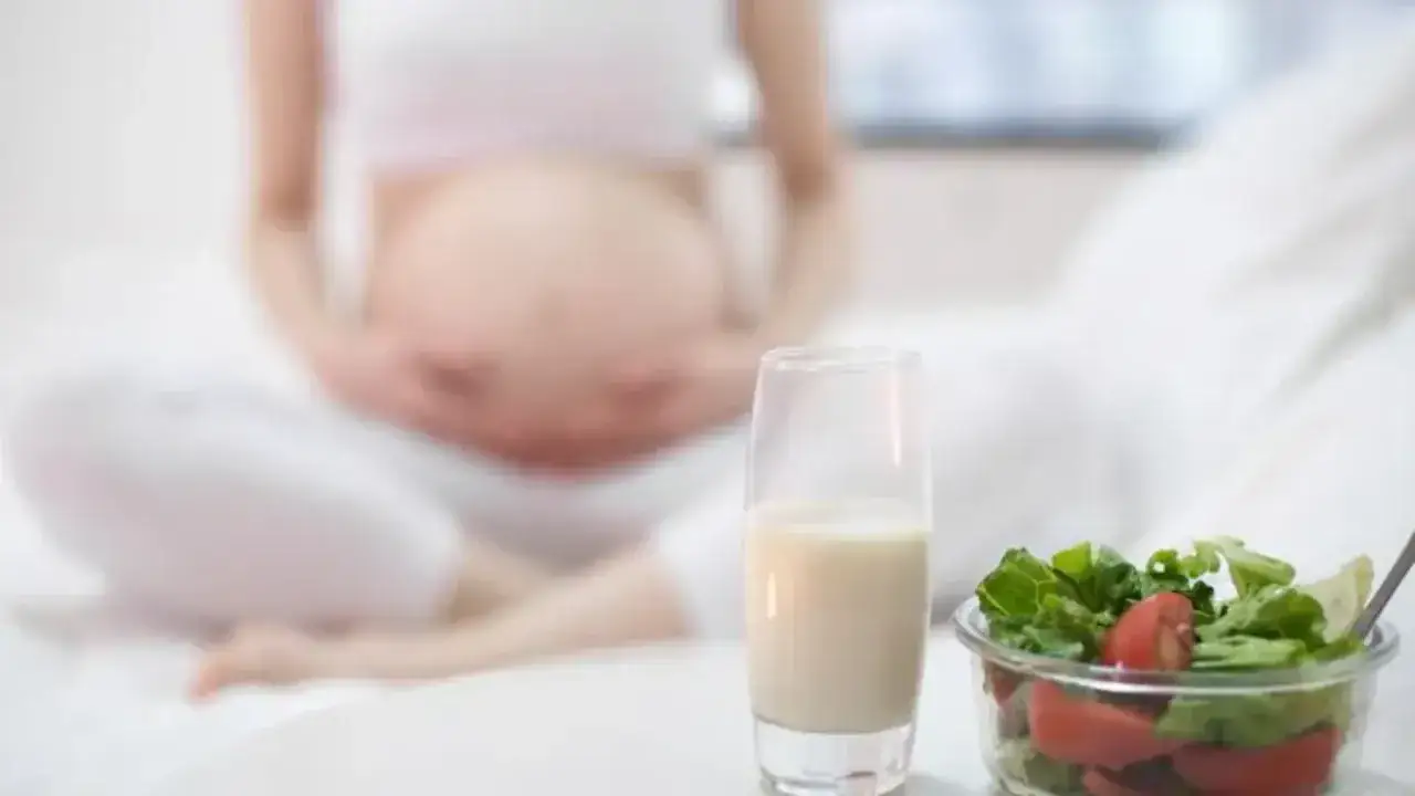 Kobieta w ciąży z brzuszkiem, obok szklanka mleka i miska sałatki. Zdrowa dieta pomaga zapobiegać zaparciom w ciąży.