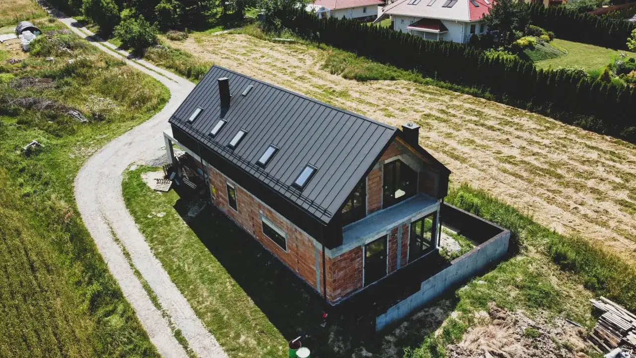 Nowy dom z cegły z czarnym dachem na rąbek, otoczony zielenią i polami.