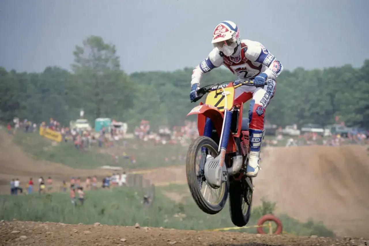 Motociclista Andre Malherbe salta en una carrera de motocross, con público al fondo.
