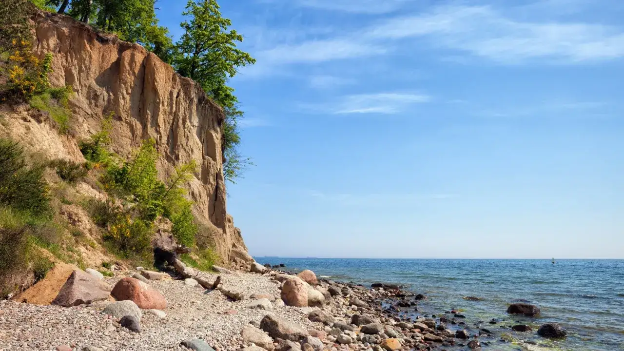 Klif Orłowski z zielenią na szczycie i kamienistą plażą u podnóża. Błękitne niebo i spokojne morze tworzą malowniczy krajobraz.