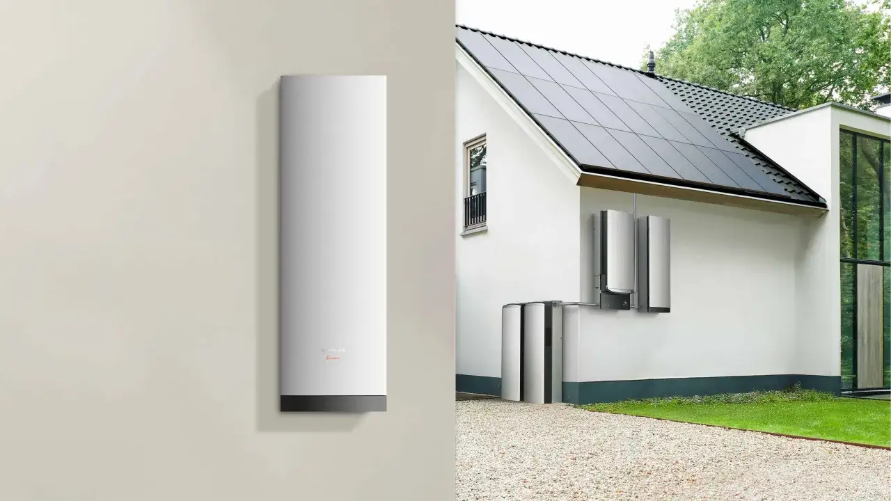 Modernt solcellsbatteri utomhus monterat p&aring; en vit v&auml;gg bredvid ett hus med solpaneler p&aring; taket.