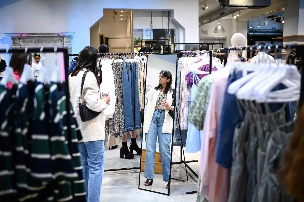 Kobiety przeglądają ubrania w sklepie z odzieżą. Wiele wieszak&oacute;w z ubraniami, typowymi dla **marki fast fashion**, tworzy tło dla sceny.