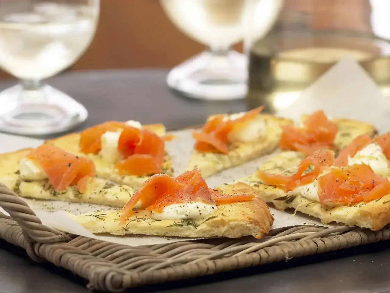Köstliche kleine Pizza-Stücke mit Lachs und Spinat, serviert auf einem Korbtablett, mit Weißwein im Hintergrund.