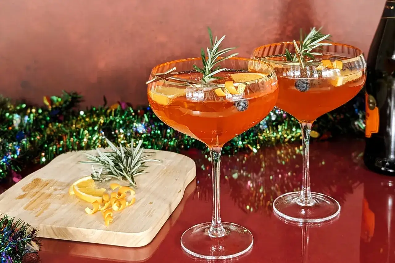 Zwei Aperol-Cocktails mit Rosmarin und Orange, bereit f&uuml;r Aperol Weihnachten. Bunte Lichterkette im Hintergrund.
