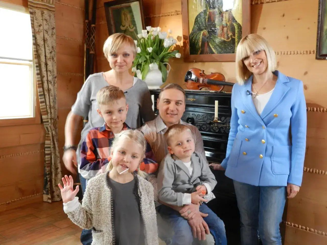 Rodzina z czwórką dzieci, w tym mała dziewczynka z lizakiem. W tle skrzypce i obrazy.