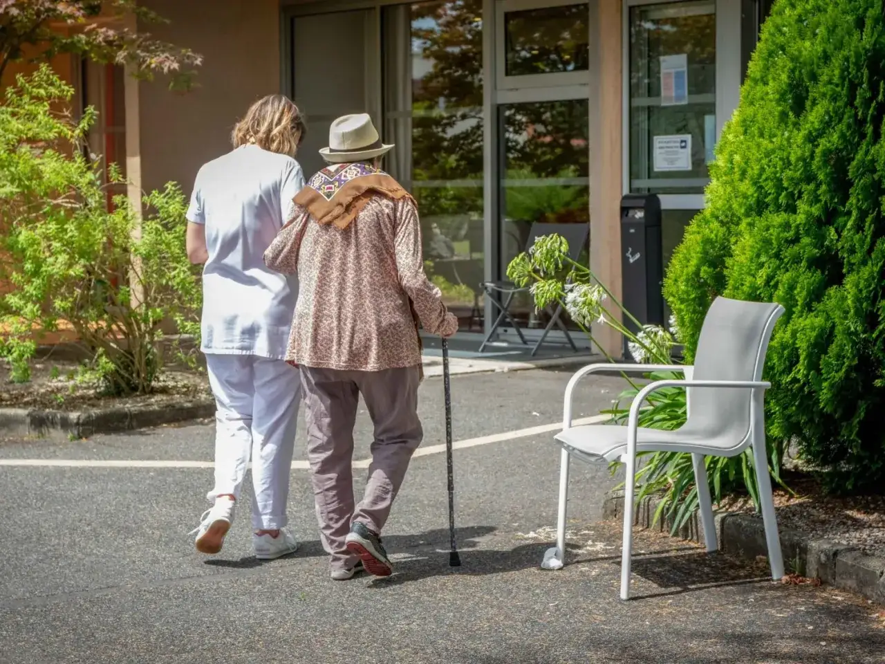 Une aide-soignante accompagne une personne âgée, canne à la main, dans le cadre d'une mise sous protection. Le soleil brille sur le chemin.