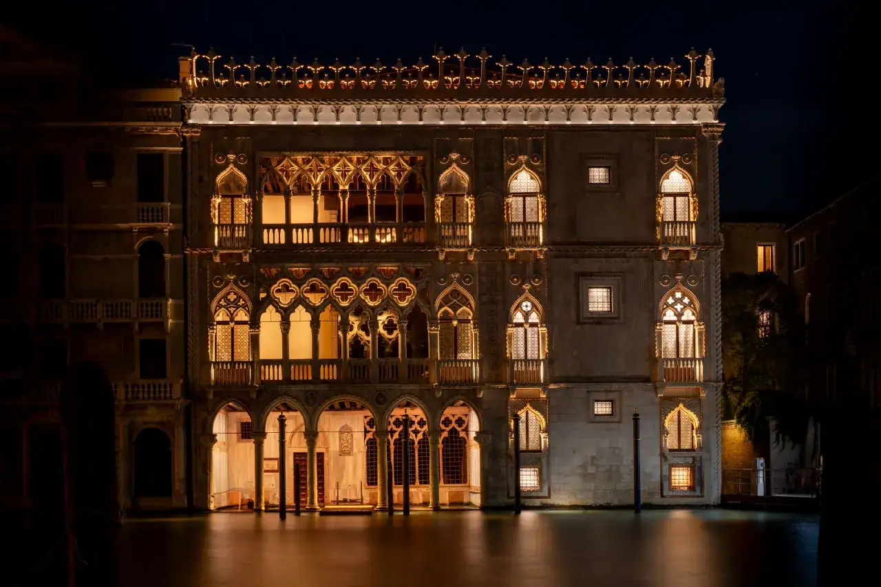 Palazzo Cadoro, illuminato di notte, si specchia sull'acqua. Le sue finestre gotiche e i dettagli architettonici brillano, creando un'atmosfera magica.