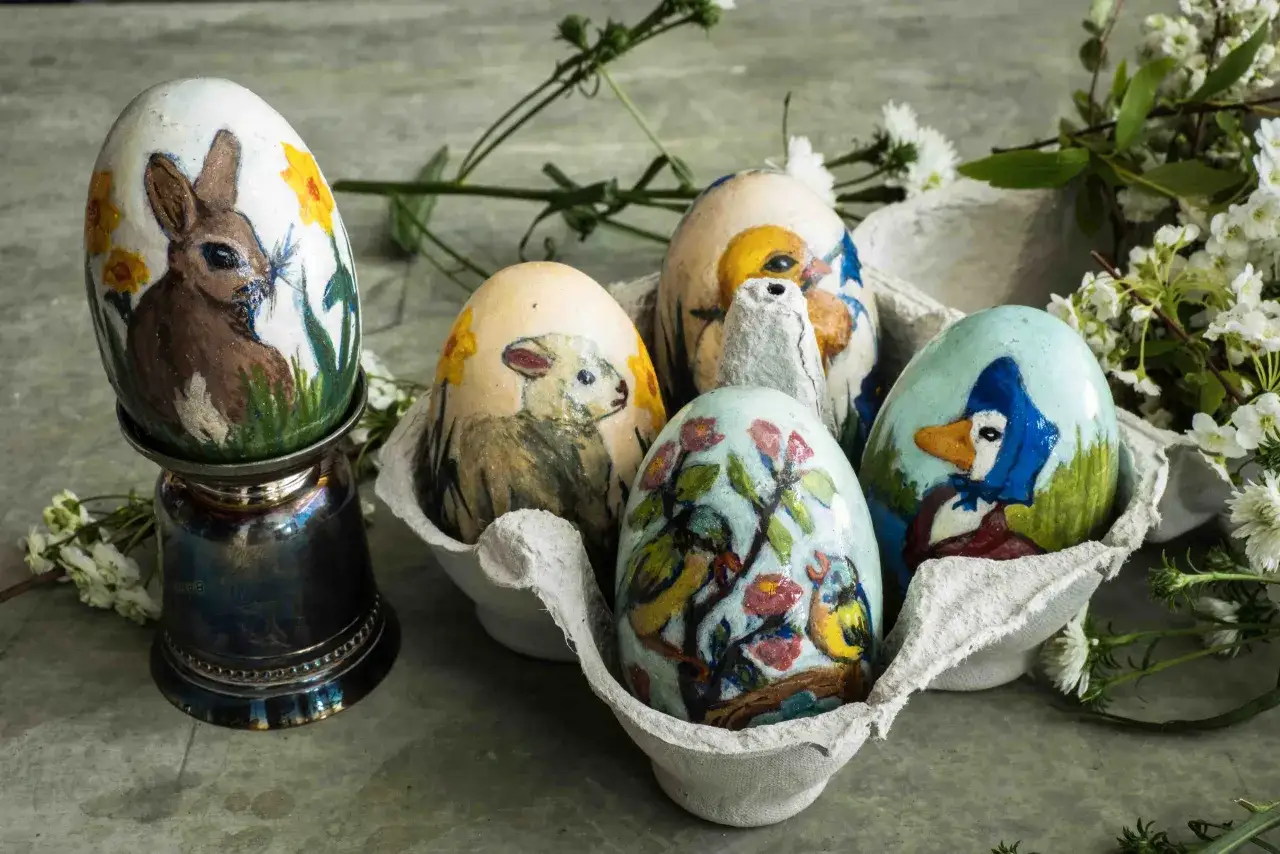 Uova di Pasqua dipinte con coniglio, agnello, uccellini e un'anatra con foulard. Le decorazioni uova di pasqua sono in un portauova di cartone, con fiori bianchi sullo sfondo.