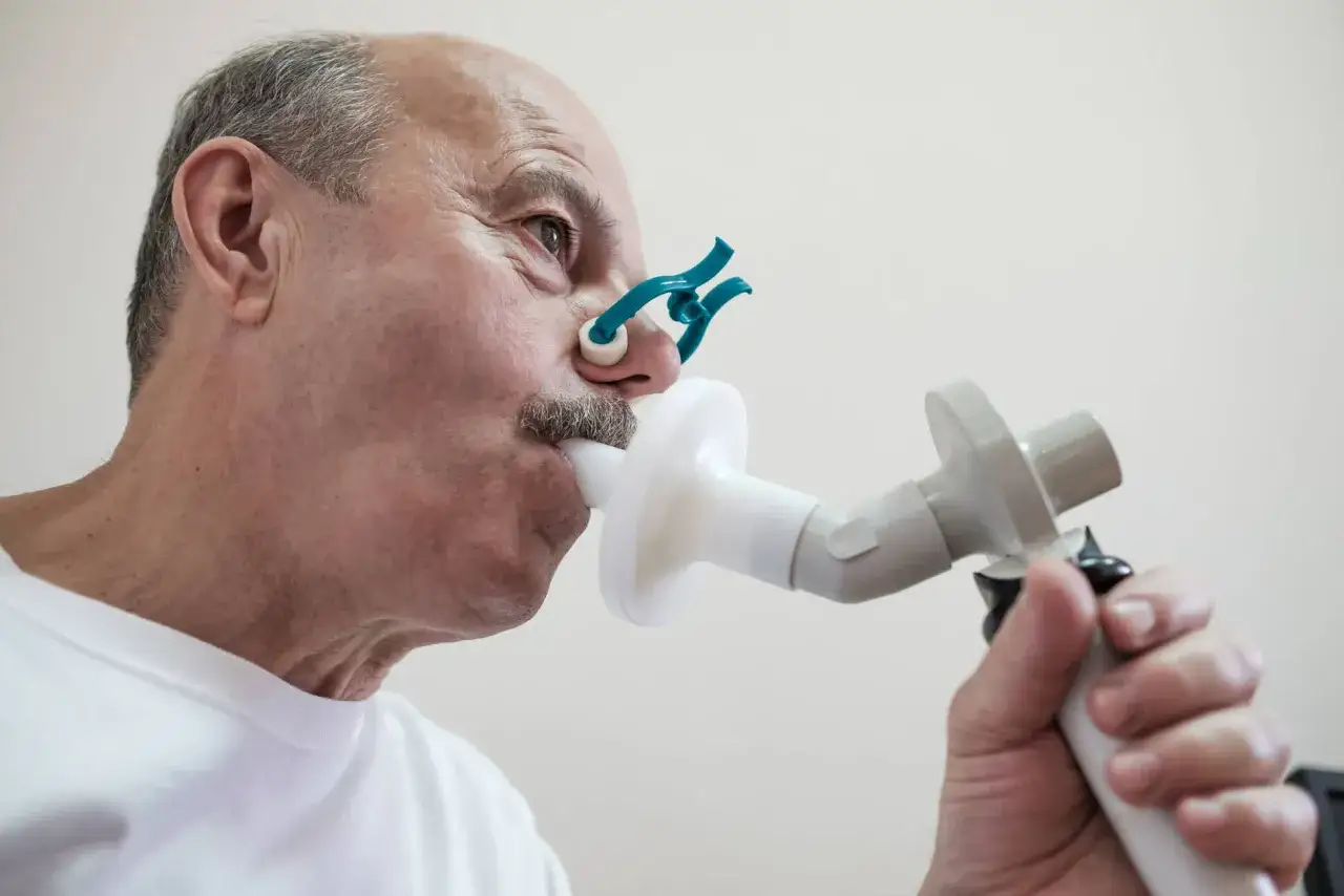 Starszy mężczyzna z klipsem na nosie wykonuje badanie spirometryczne. Długość życia z POChP zależy od wielu czynników.