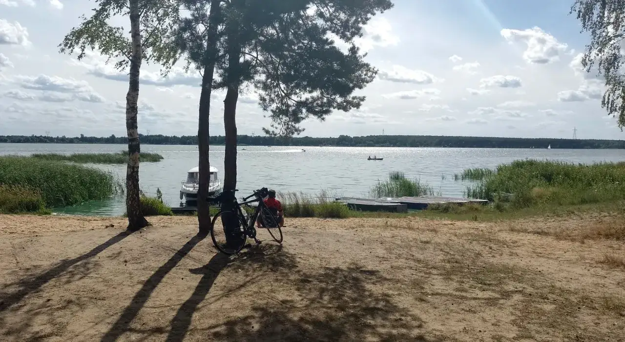 Spokojny dzień nad jeziorem w Poraju. Rower i osoba odpoczywają na piaszczystej plaży, w tle widać łodzie i żaglówki.