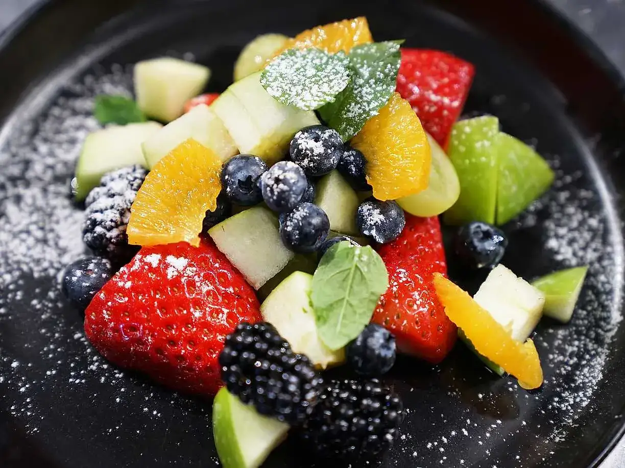 En färgstark ensalada de frutas med jordgubbar, blåbär, björnbär, äpple och apelsin, pudrad med florsocker.