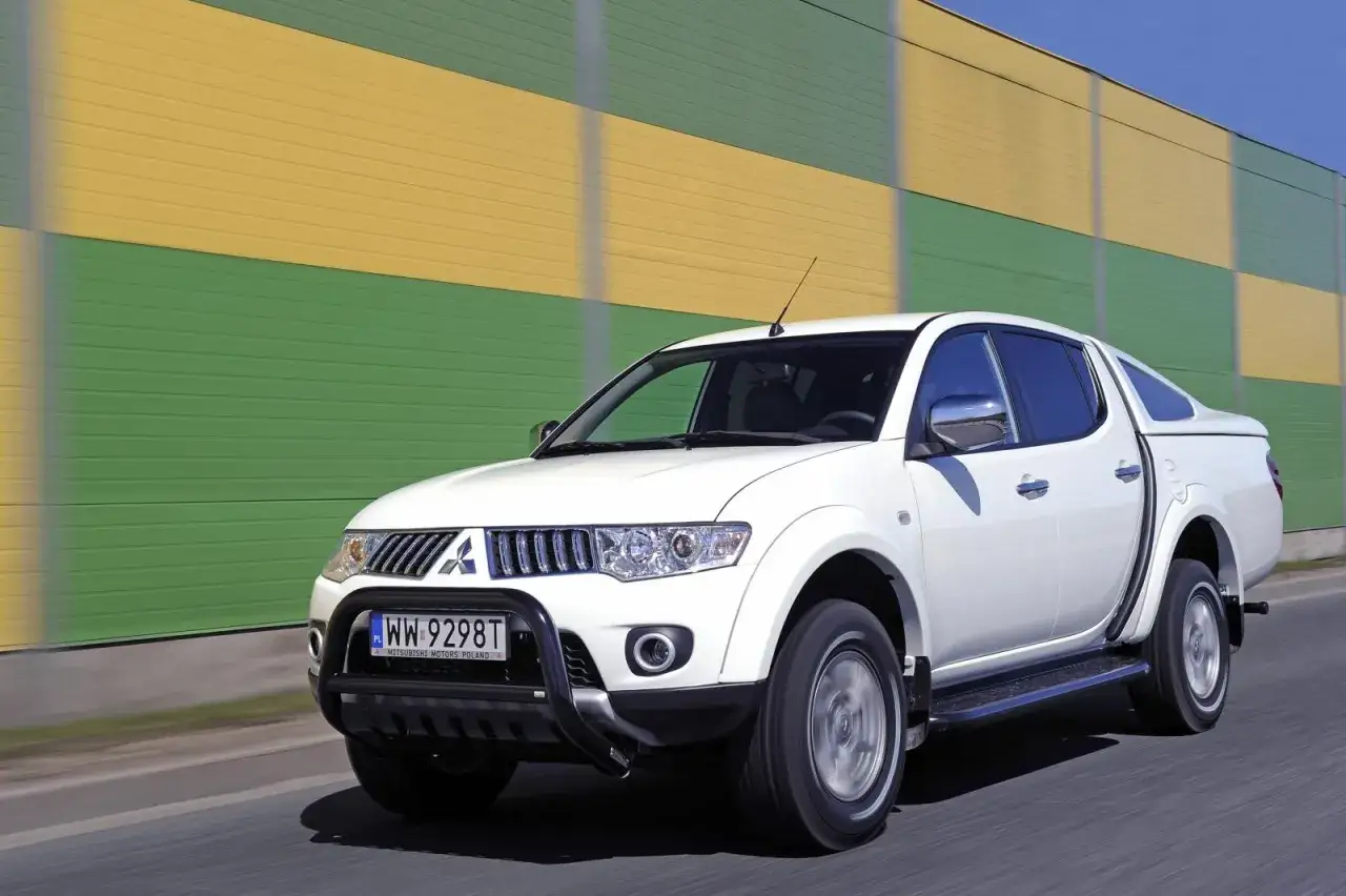 Biały model terenówki Mitsubishi L200 z czarną osłoną zderzaka i nakładką na pakę, pędzi po drodze na tle zielono-żółtego ekranu dźwiękochłonnego.