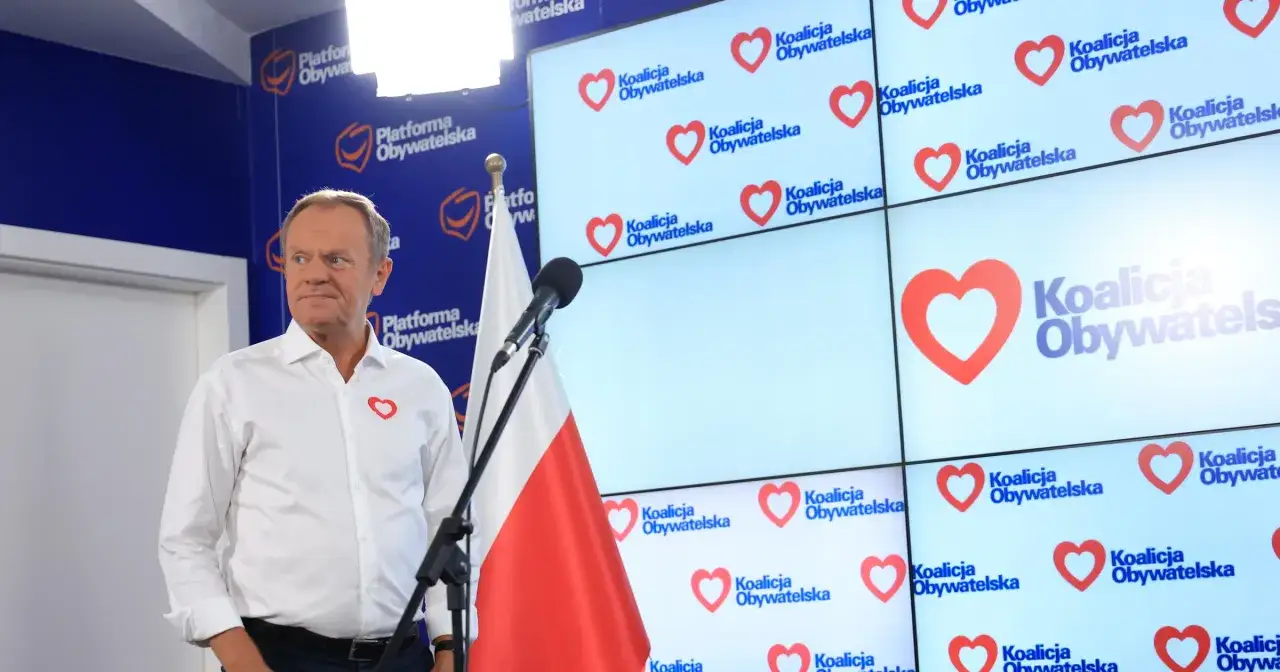 Donald Tusk w białej koszuli z sercem, na tle logo Koalicji Obywatelskiej i flagi Polski.