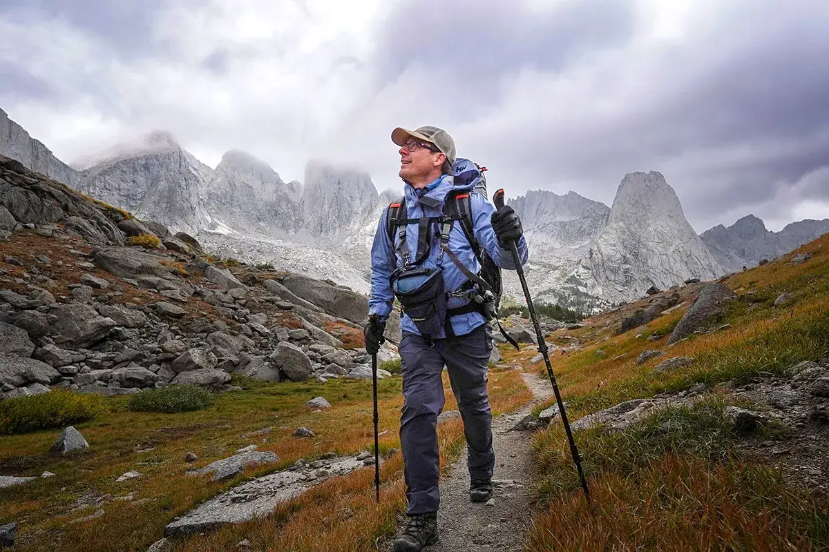 Mężczyzna w czapce i kurtce z plecakiem idzie ścieżką w górach, używając kijków. To idealne miejsce na speed hiking.
