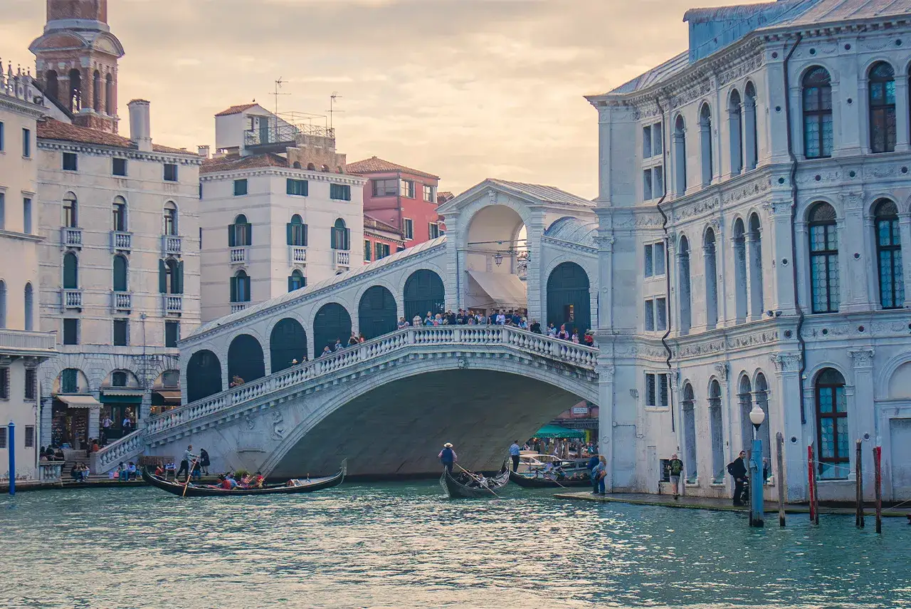 Gondole scivolano sull'acqua sotto il maestoso ponte di Rialto a Venezia, con turisti che ammirano il panorama.