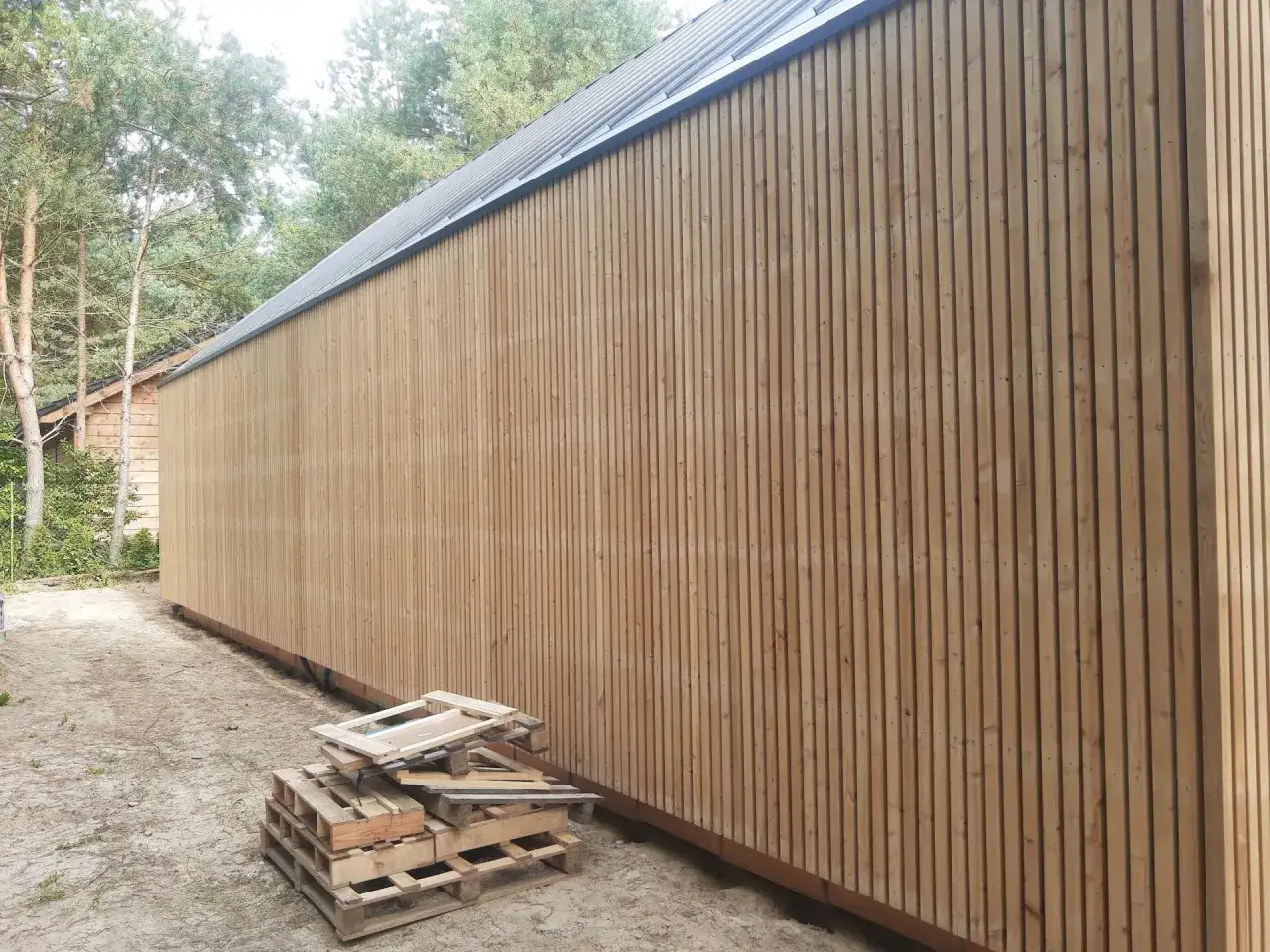 Nowa elewacja z drewna, tworząca efekt deski, otoczona sosnami. Obok palety i resztki materiałów budowlanych.