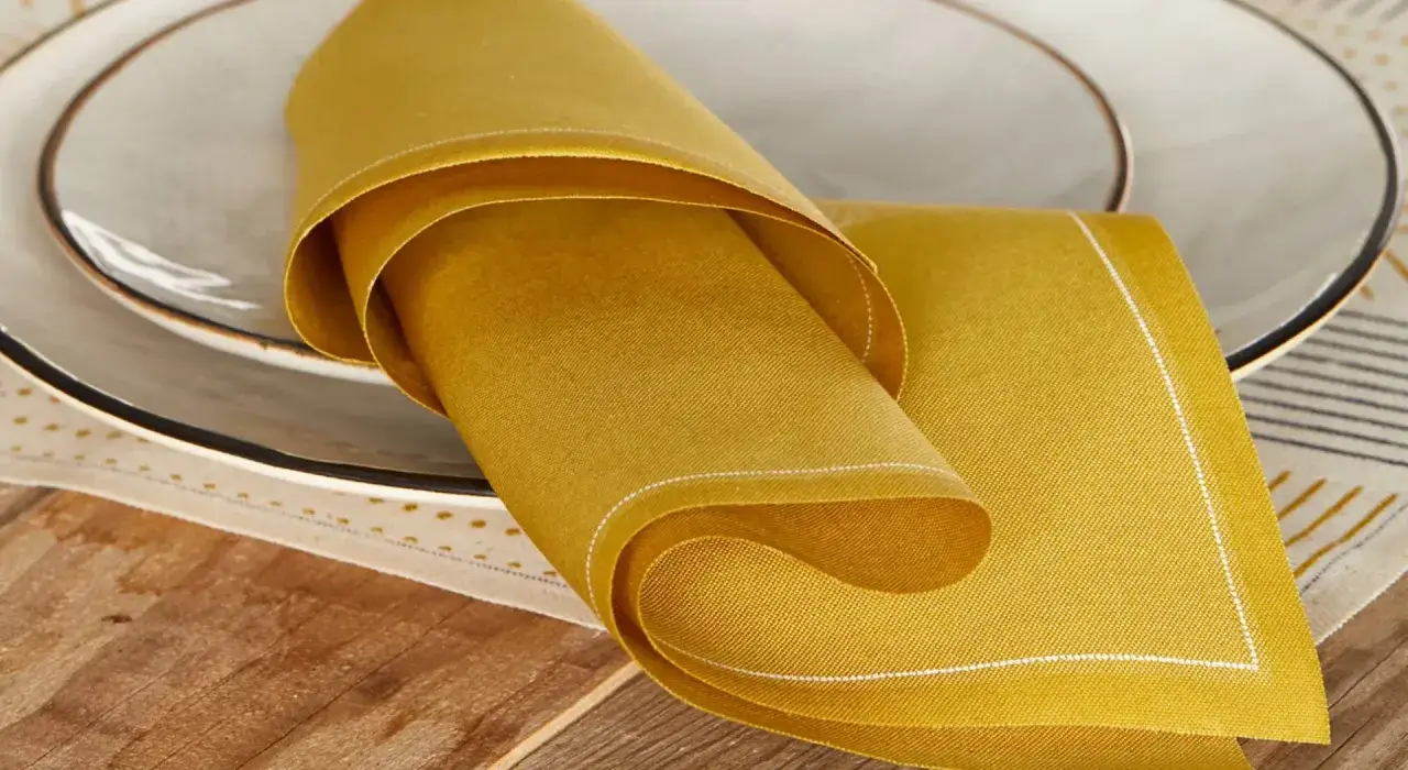 Serviette de table en lin ocre, pliée sur une assiette. Un choix idéal pour savoir quel tissu pour serviette de table.
