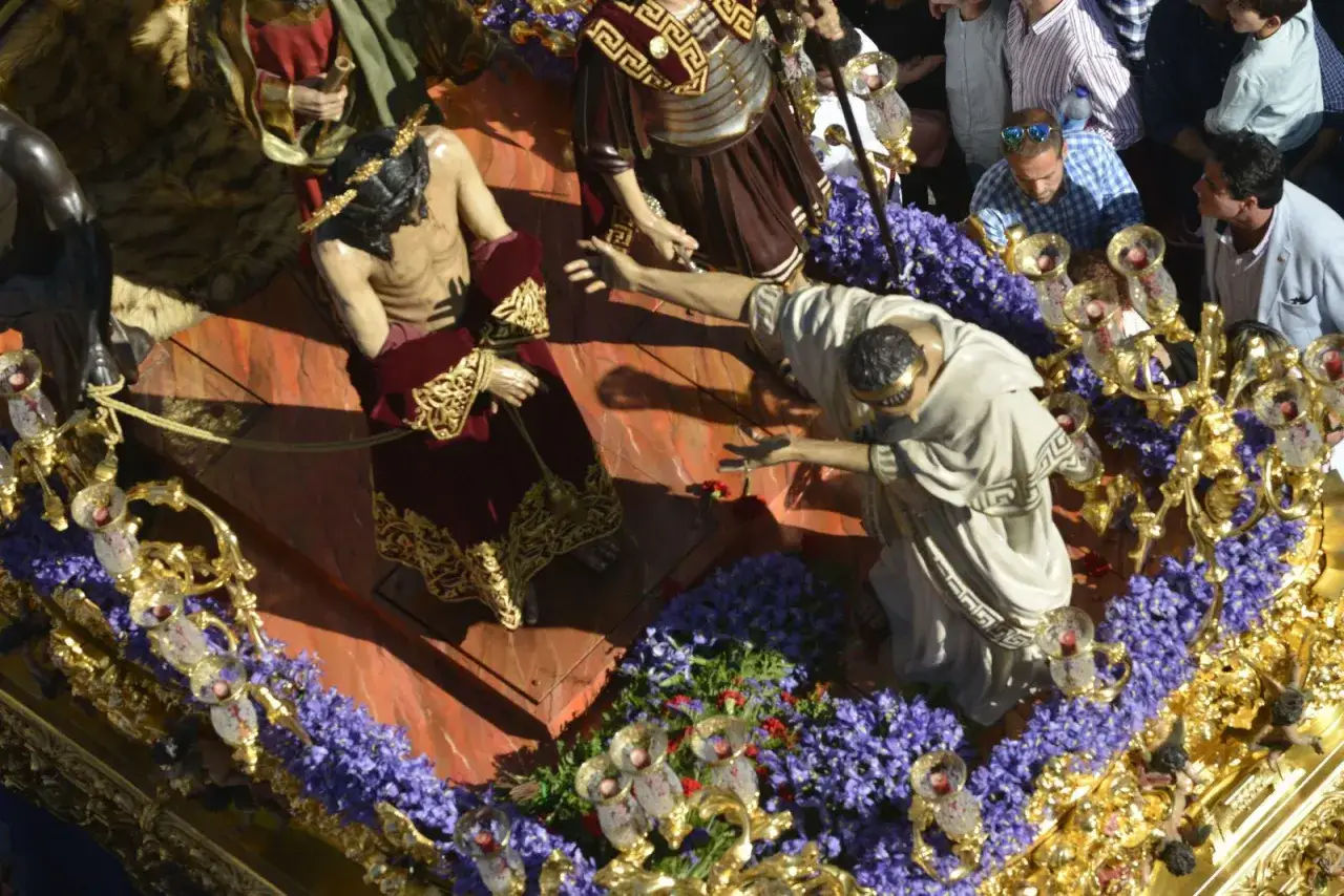 Detalle de **procesiones martes santo**: Cristo con corona de espinas y un soldado romano. Flores moradas y dorados adornan el paso.