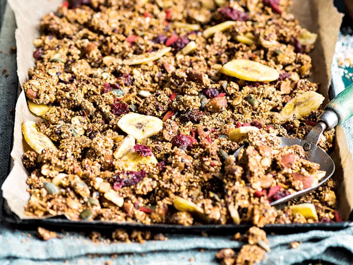 Knuspriges Granola mit Amaranth, Bananenchips und Cranberries auf Backpapier. Ein Löffel nimmt eine Portion auf.