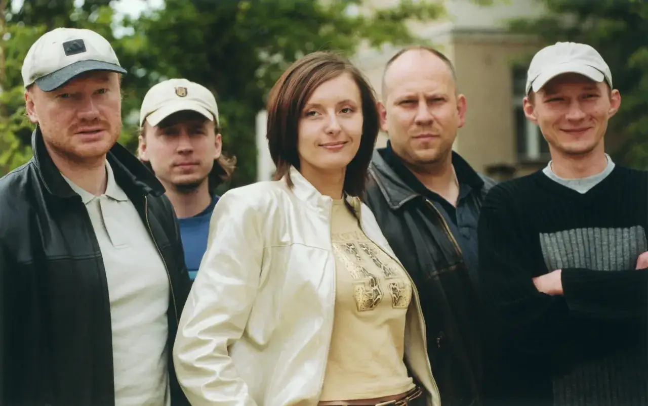 Grupa osób, w tym kobieta w błyszczącej kurtce, pozuje do zdjęcia. W tle drzewa i budynek. Wszyscy wyglądają jakby mieli coś wspólnego, może to zespół Varius Manx?