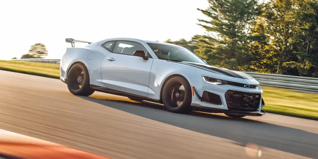 Białe Camaro ZL1 z czarnymi pasami na masce pędzi po torze wyścigowym, otoczone zielenią drzew.