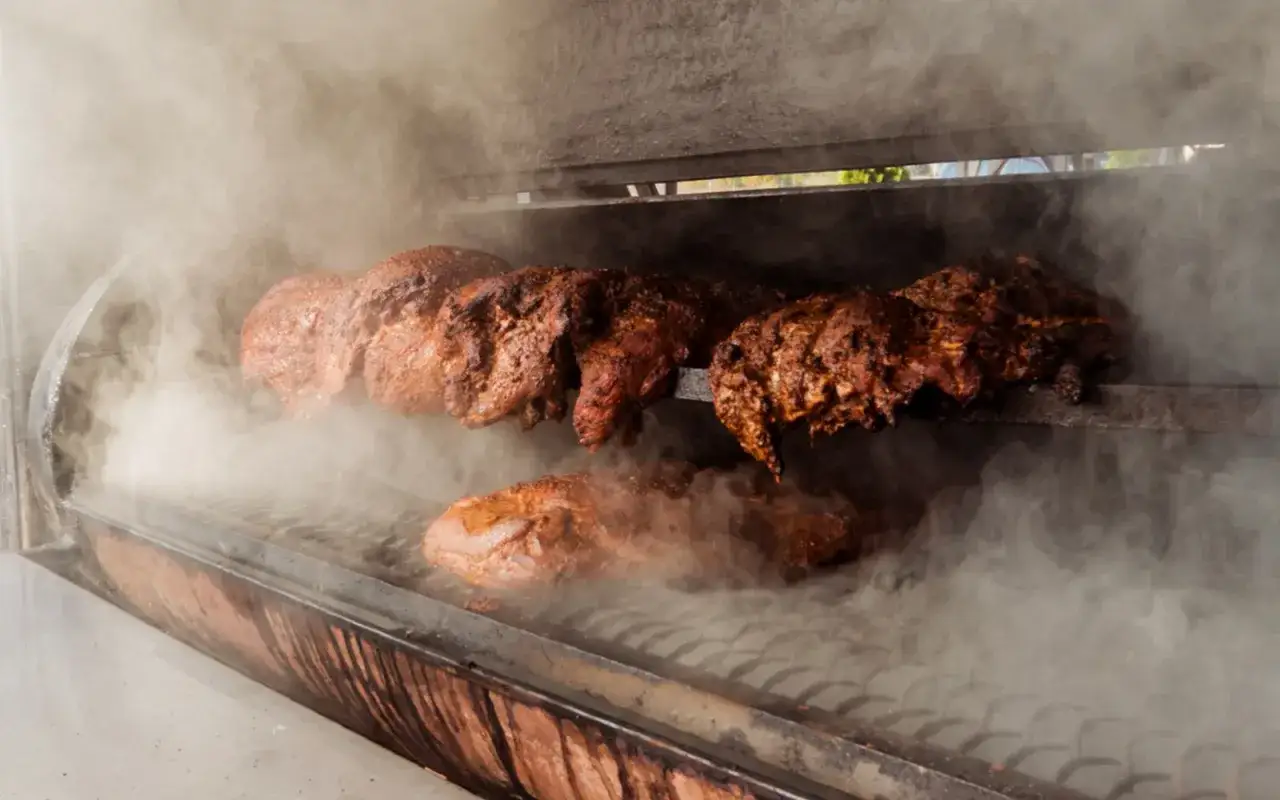 Viande rôtie sur une broche dans un fumoir, le fumage à chaud libérant une épaisse fumée.