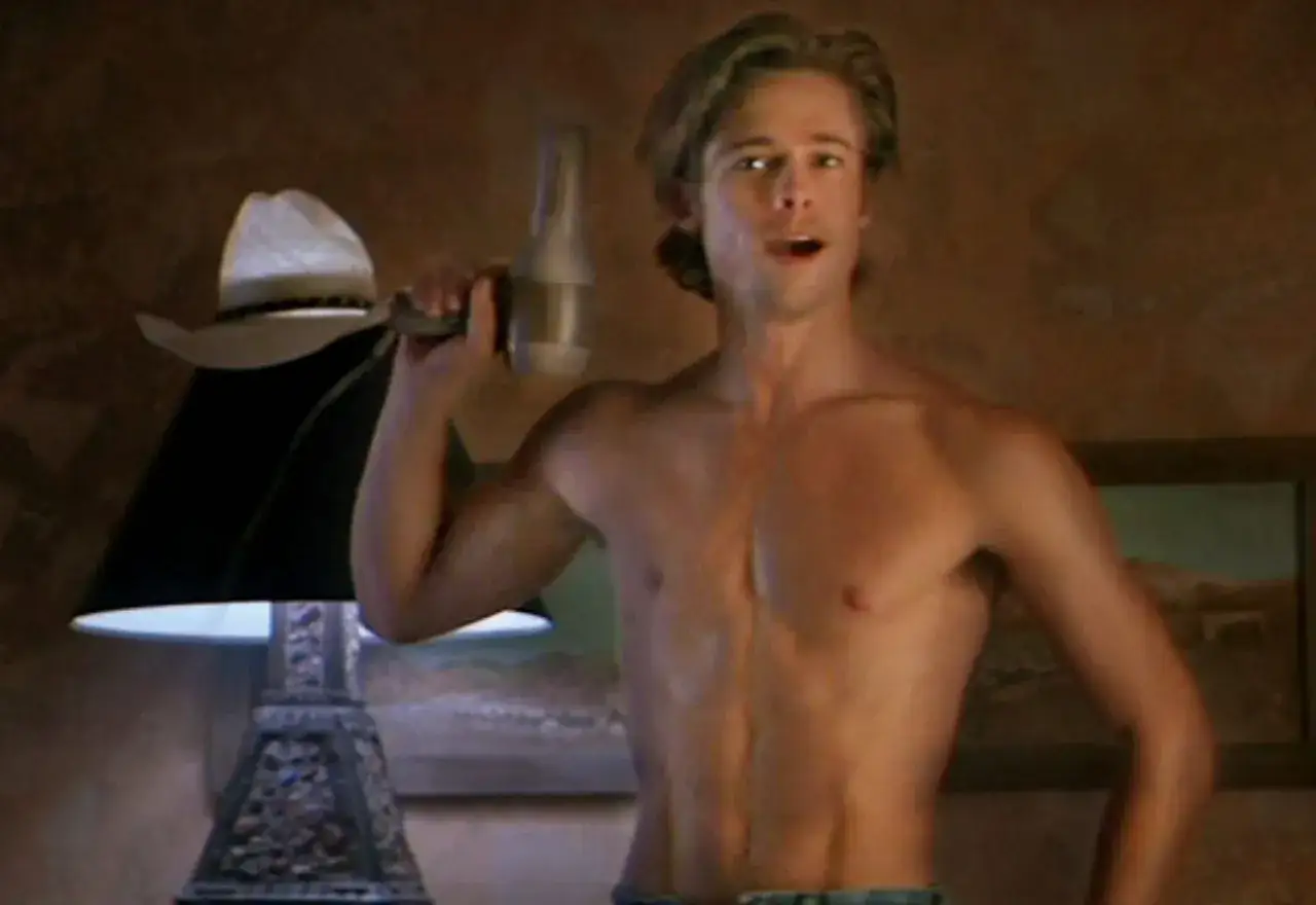 Brad Pitt ung med hårfön i handen, redo att styla sitt blonda hår. En cowboyhatt vilar på en lampa.