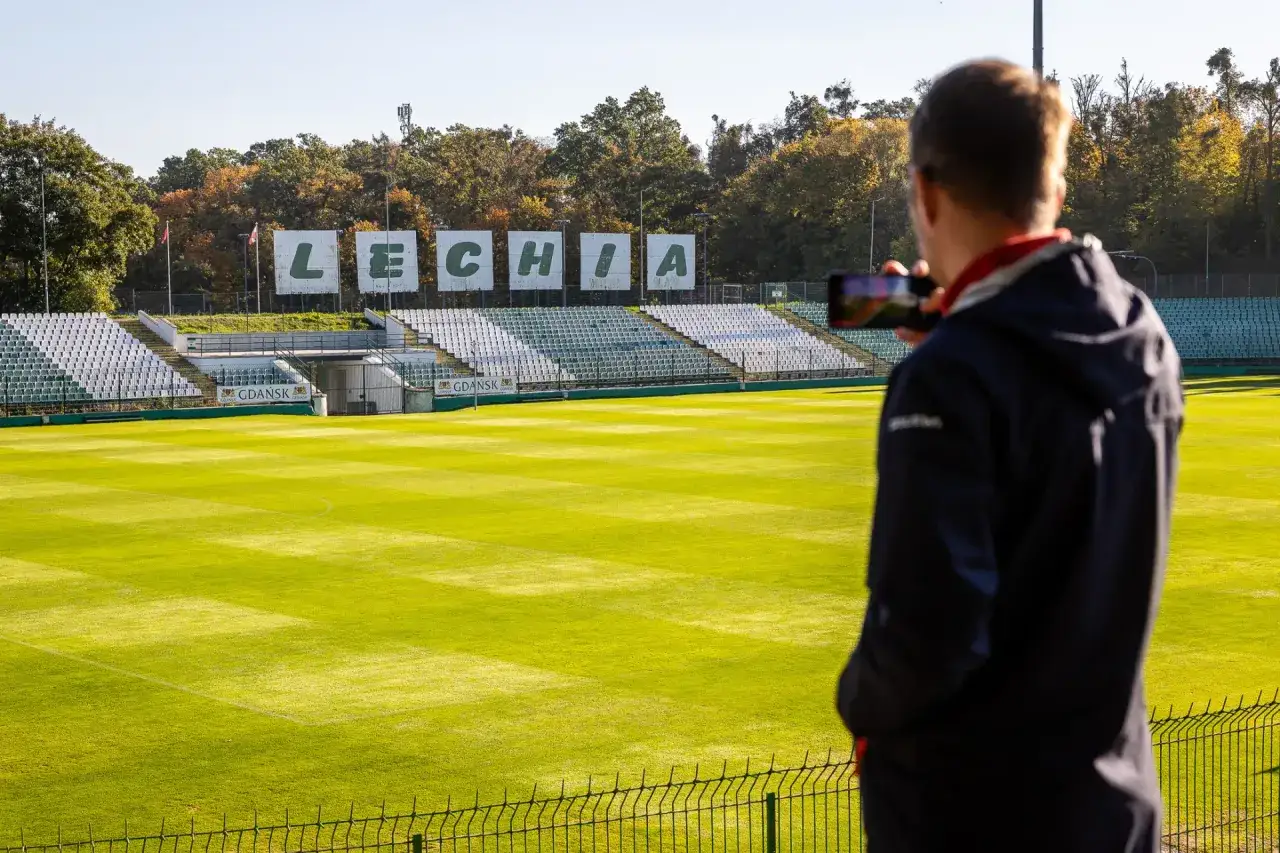 Widok na stadion Lechii Gdańsk. Mężczyzna z telefonem w ręku podziwia boisko, myśląc o przyszłych meczach piłkarzy z Gdańska.