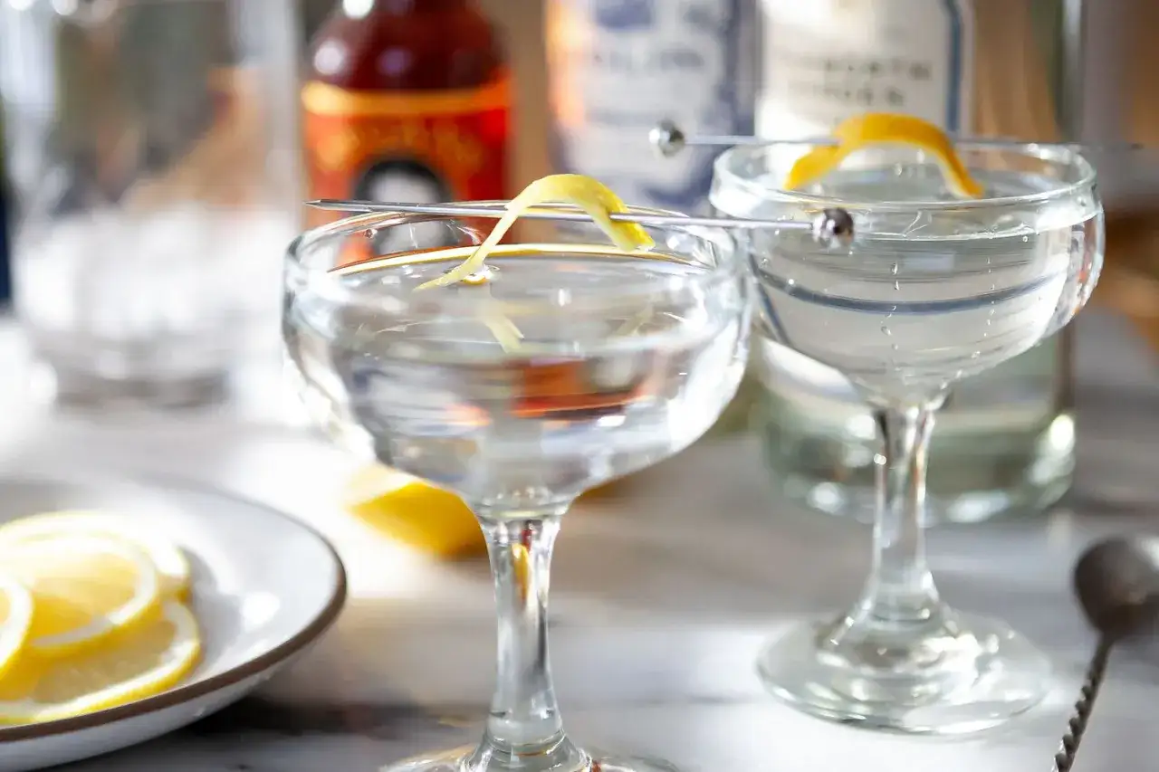 Klasyczny gin martini przepis: dwa eleganckie kieliszki z orzeźwiającym drinkiem, ozdobione skórką cytryny. Obok talerzyk z plasterkami cytryny.