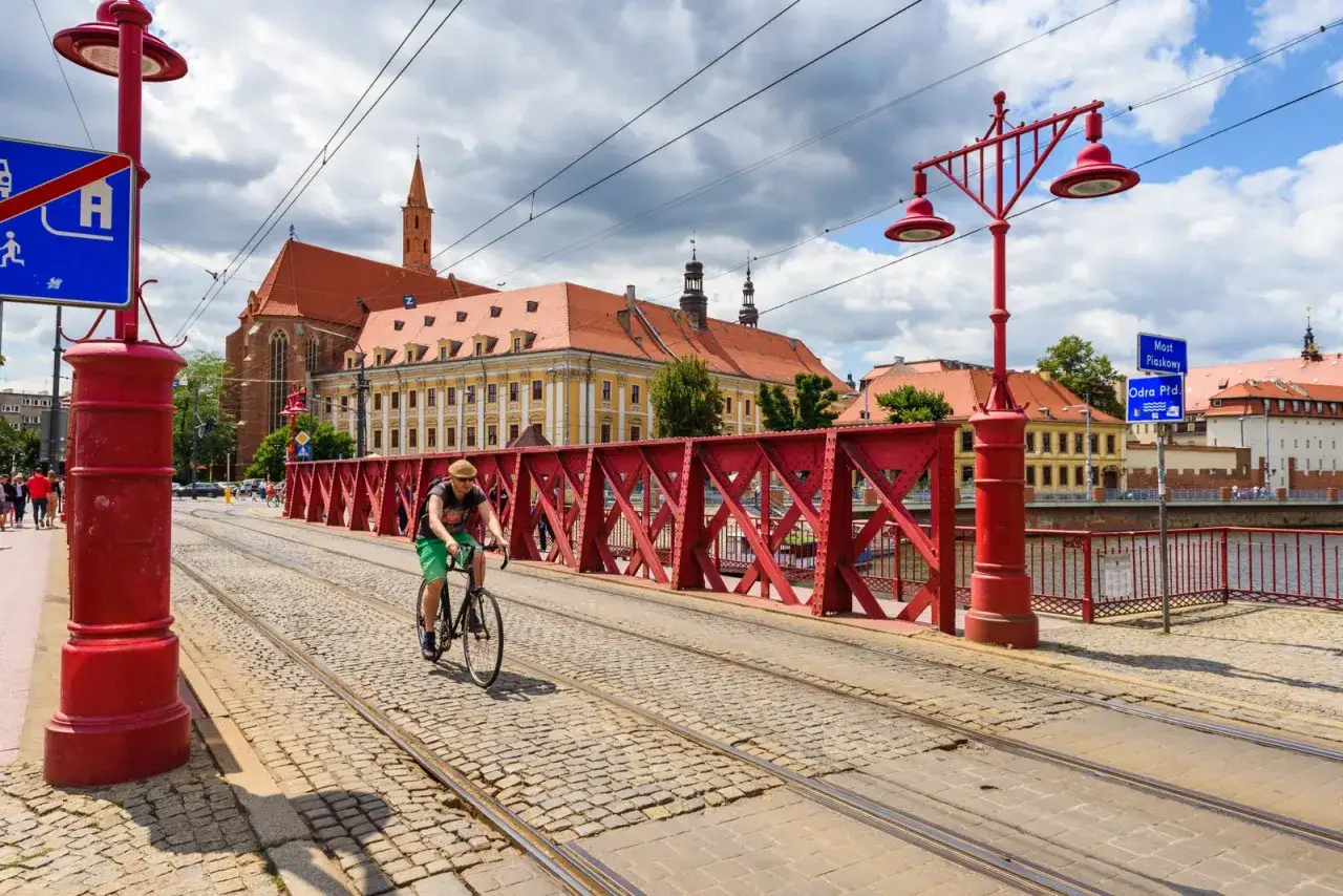 Mężczyzna na rowerze jedzie po brukowanej drodze rowerowej z torami tramwajowymi, w tle zabytkowe budynki i czerwony most.