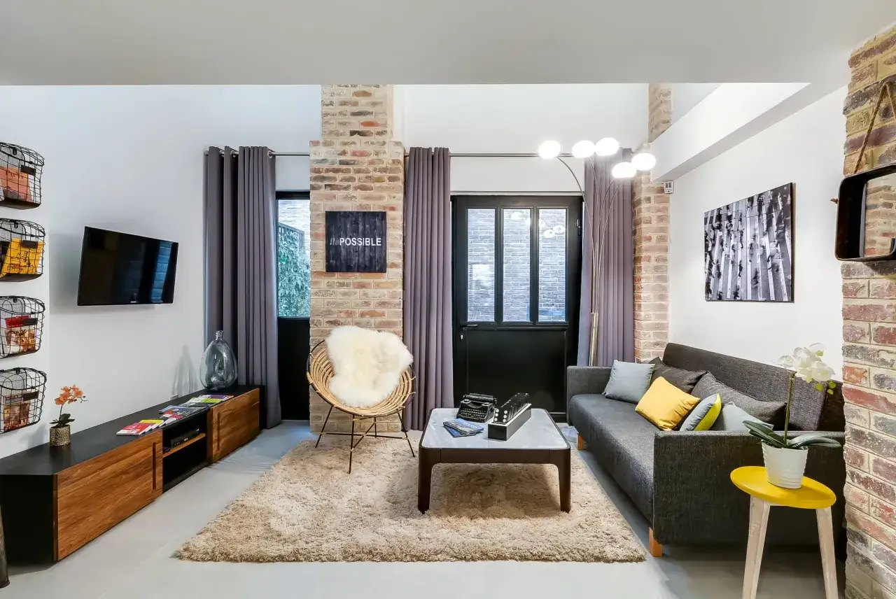 Salon moderne avec murs en briques apparentes, canapé gris, fauteuil en rotin et rideaux de type pli rideau.