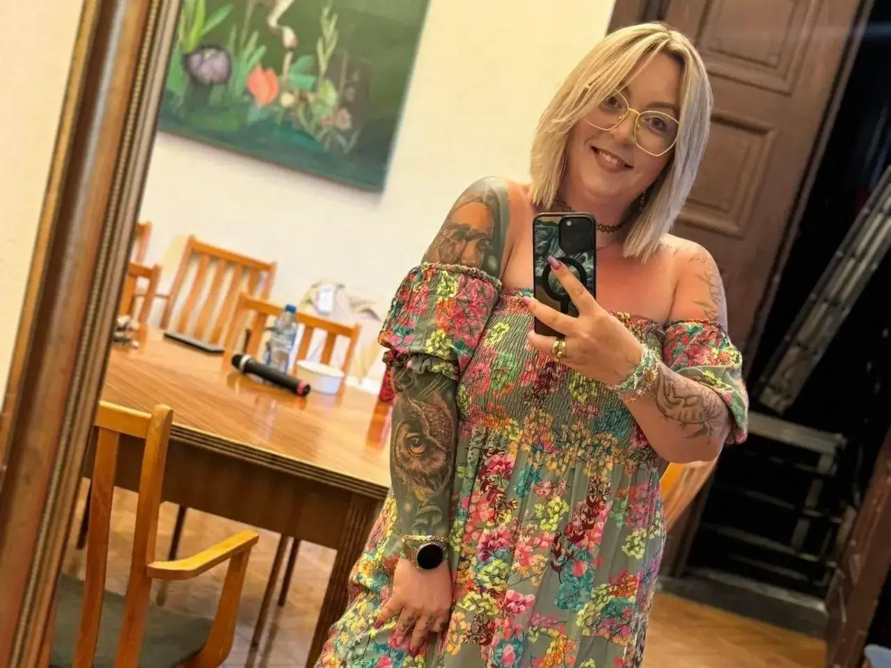 Uśmiechnięta mama na obrotach, w kwiecistej sukience i z tatuażami, robi sobie selfie w lustrze.