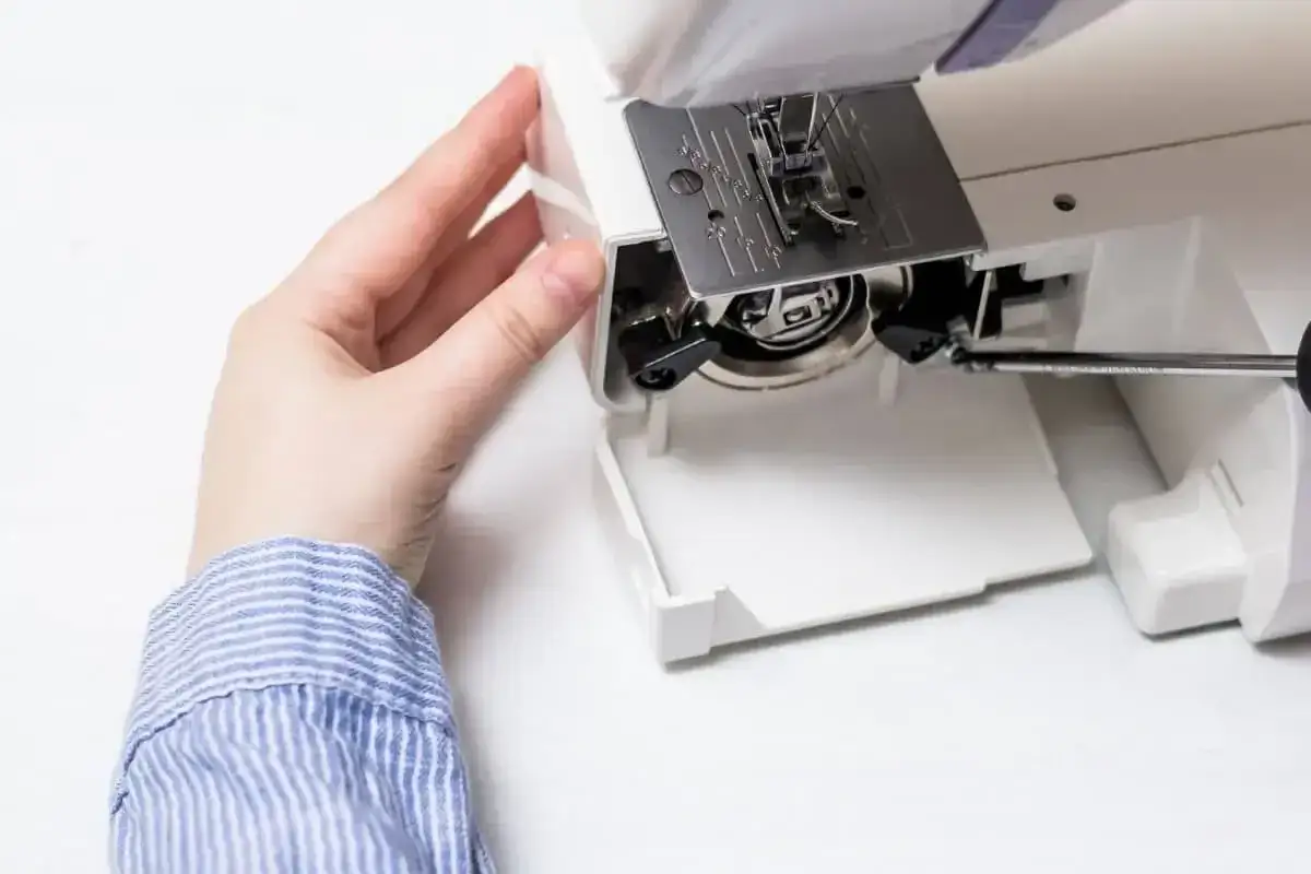 Dłoń zdejmuje osłonę maszyny do szycia, odsłaniając mechanizm bębenka. Dowiedz się, jak oliwić maszynę do szycia, by działała płynnie.