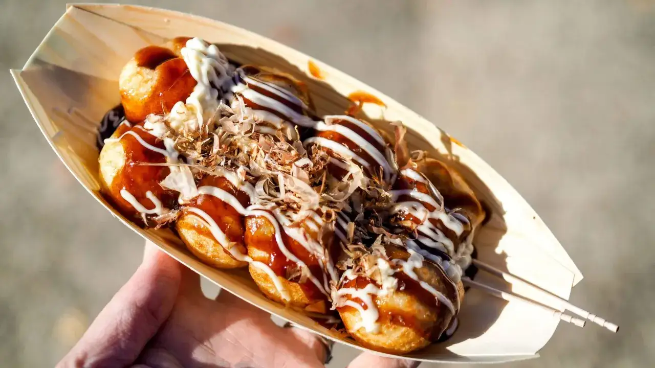 A hand holds a wooden boat filled with delicious takoyaki food, drizzled with sauce and mayonnaise, and topped with bonito flakes.