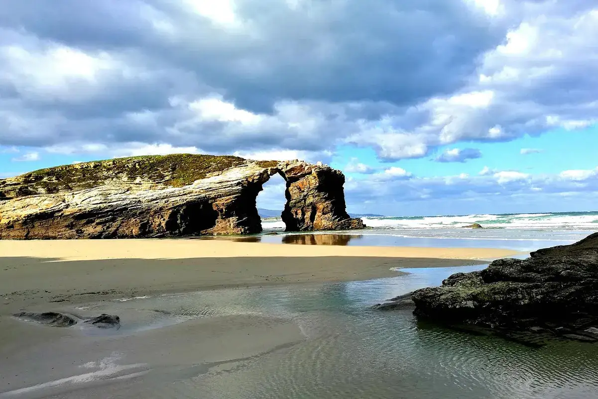 Arco natural en la playa de las Catedrales, Lugo. El mar Cantábrico besa la arena dorada bajo un cielo nublado.