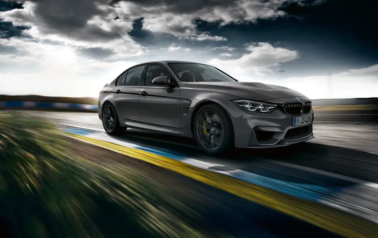 Szare BMW M3 F80 pędzi po torze wyścigowym, chmury na niebie dodają dramatyzmu.