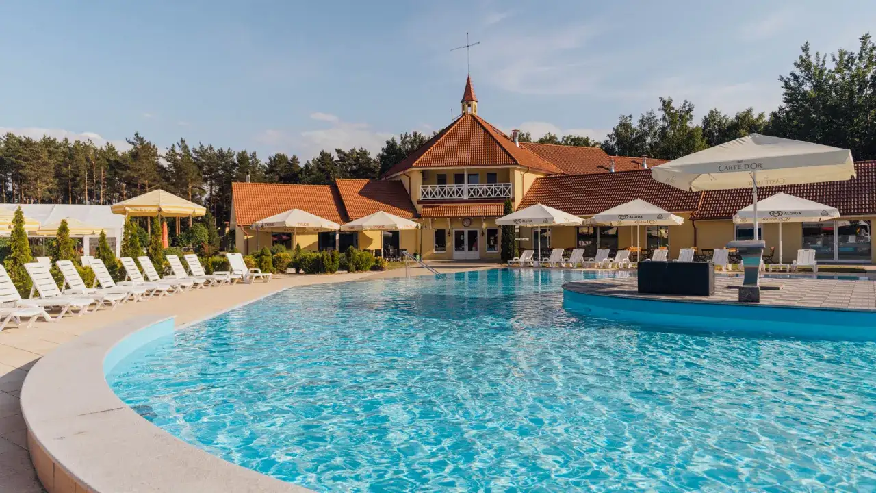 Piękny, błękitny basen z leżakami i parasolami, a w tle żółty budynek z czerwoną dachem. Idealne miejsce na wypoczynek, jak domek przy samej plaży nad morzem.