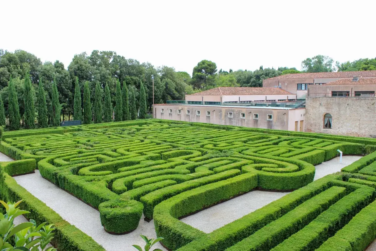 Un intricato labirinto di siepi verdi a Venezia, con un edificio moderno sullo sfondo e alberi alti.