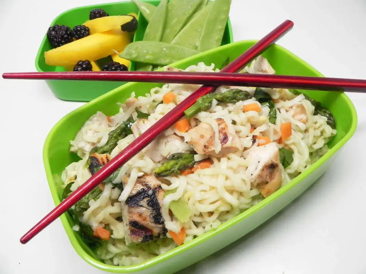 A vibrant noodle bento box filled with chicken, asparagus, and carrots, accompanied by a side of mango, blackberries, and snap peas.