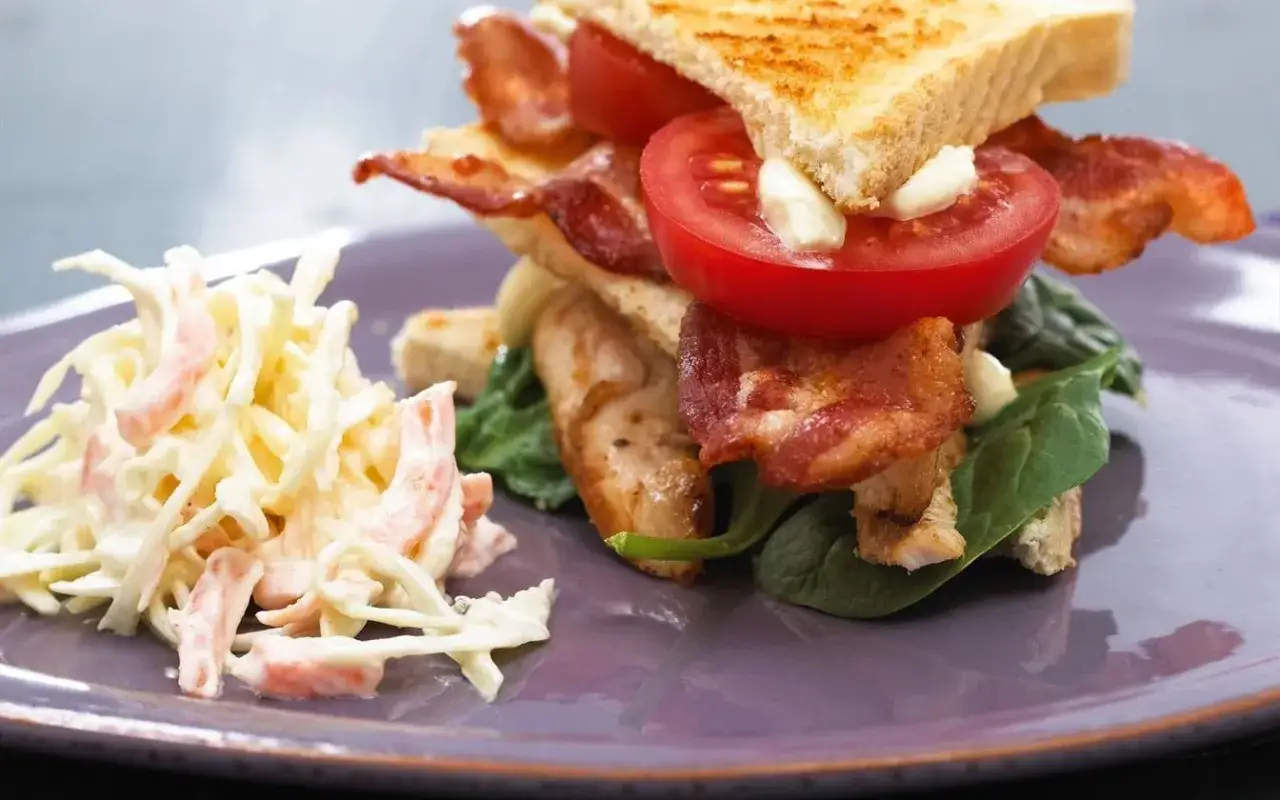 Ein saftiger Club Sandwich mit knusprigem Speck, Tomaten und H&auml;hnchen, serviert mit einem cremigen Krautsalat.