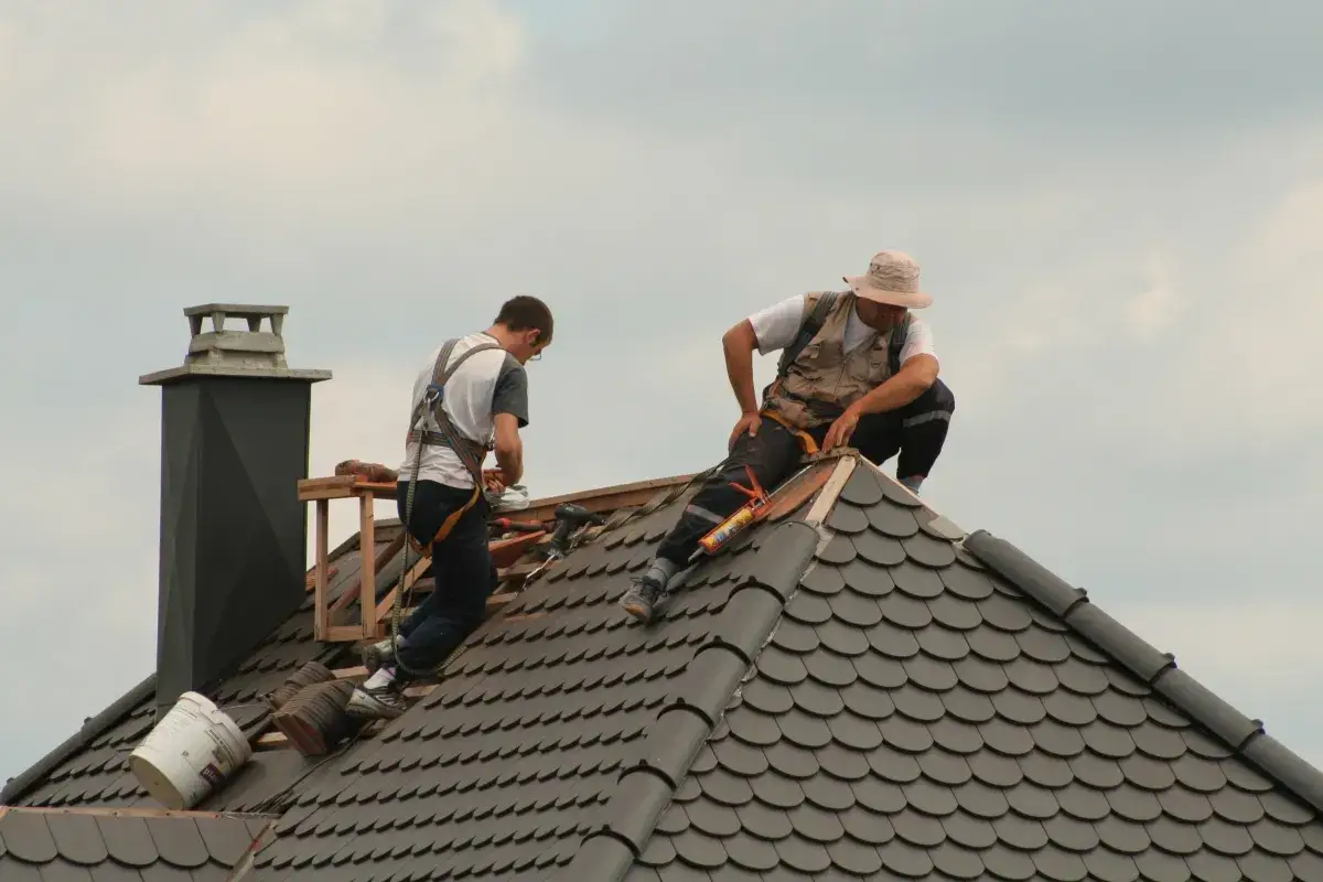 Dw&oacute;ch dekarzy wykonuje pracę na dachu. Jeden z nich używa pistoletu z uszczelniaczem, drugi przygotowuje materiały.