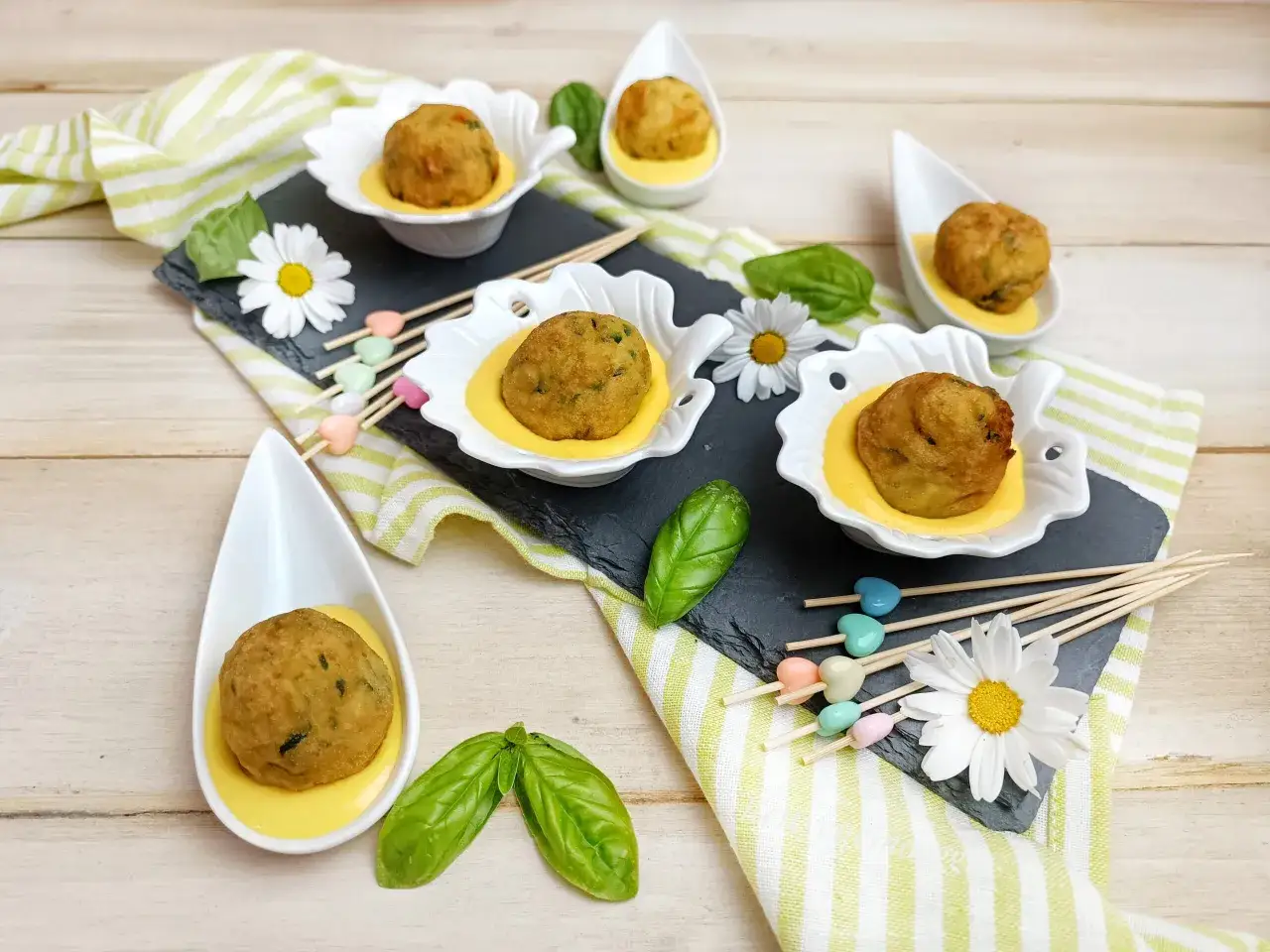 Deliziose polpette di zucchine dorate, servite in eleganti ciotoline con una salsa gialla, guarnite con basilico fresco e fiori di margherita.