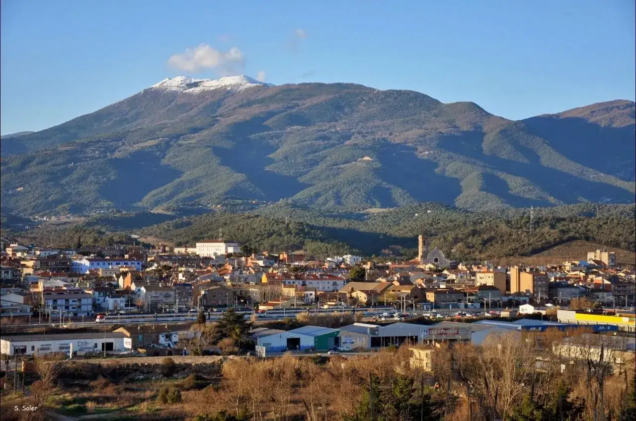 Vista de un pueblo con un turo del home nevado al fondo. El sol ilumina las casas y la vegetaci&oacute;n.