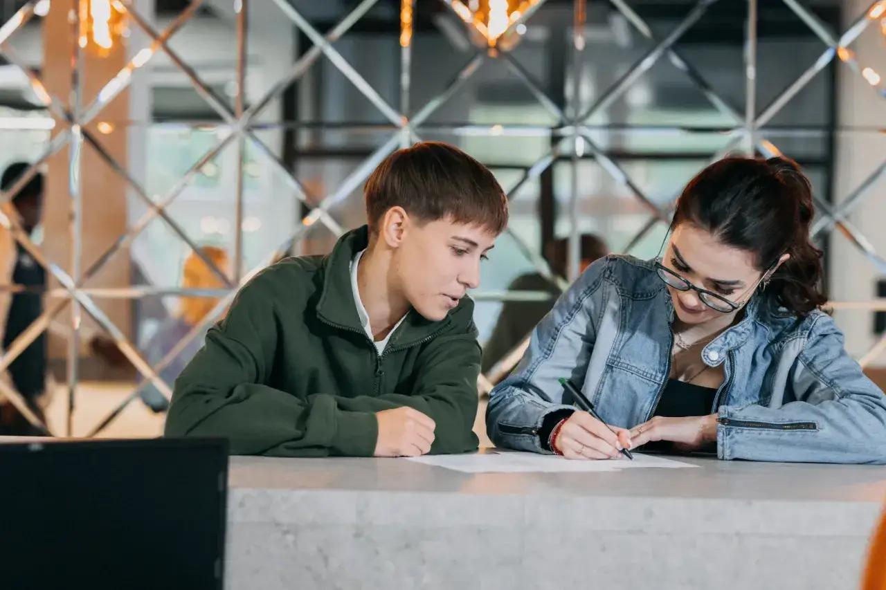 Dwoje studentów przy biurku, jeden pisze, drugi patrzy. Wspólnie pracują nad wnioskiem o stypendium socjalne.